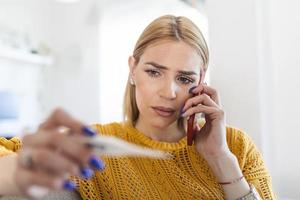 jong vrouw is op zoek Bij de thermometer. ze heeft koorts, roeping haar dokter. mooi vrouw met symptomen gebeld covid-19 noodgeval aantal naar verslag doen van haar medisch voorwaarden foto