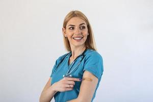 portret van een vrouw dokter glimlachen na krijgen een vaccin. medisch arbeider tonen haar arm met verband na ontvangen vaccinatie. foto