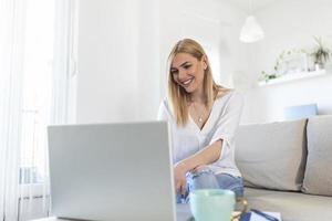 positief jong vrouw freelancer aan het kijken video in blog Aan netbook en hebben een koffie Bij comfortabel huis. mooi jong bedrijf vrouw werken Aan laptop.freelancer Verbinden naar internet foto