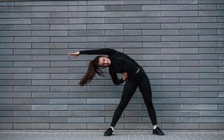 jong sportief meisje in zwart sportkleding buitenshuis aan het doen uitrekken in de buurt grijs muur foto