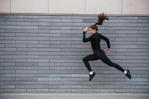 jong sportief meisje in zwart sportkleding rennen buitenshuis in de buurt grijs muur foto