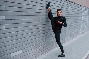 jong sportief meisje met telefoon in zwart sportkleding aan het doen poten uitrekken buitenshuis door gebruik makend van grijs muur foto