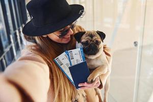 jong vrouw passagier in warm kleren Holding kaartjes en schattig hond in handen in luchthaven hal foto