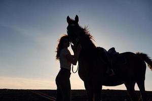 jong vrouw staand met haar paard in landbouw veld- Bij zonnig dag foto