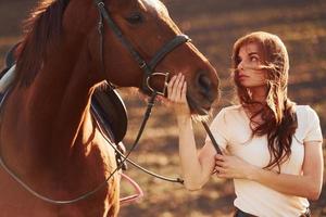 jong vrouw staand met haar paard in landbouw veld- Bij zonnig dag foto