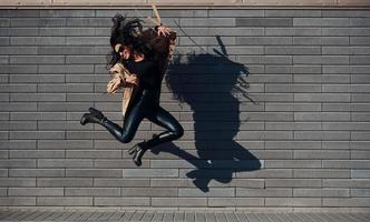 mooi brunette met gekruld haar- en in zwart kleren jumping en hebben pret buitenshuis in de buurt muur foto