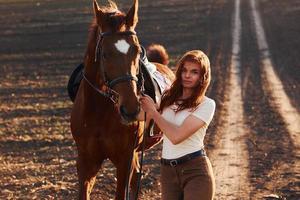 jong vrouw in beschermend hoed wandelen met haar paard in landbouw veld- Bij zonnig dag foto