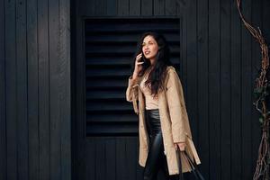 vrouw met zwart gekruld haar- staand tegen zwart houten gebouw buitenkant en gebruik makend van telefoon foto