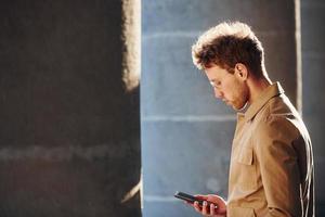 mooi hoor zonneschijn. Holding telefoon in hand. elegant jong Mens in formeel classy kleren buitenshuis in de stad foto