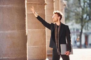 tonen gebaar door hand. met laptop in handen. elegant jong Mens in bril adn in formeel classy kleren buitenshuis in de stad foto