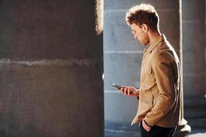 mooi hoor zonneschijn. Holding telefoon in hand. elegant jong Mens in formeel classy kleren buitenshuis in de stad foto