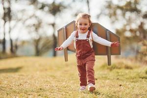 schattig weinig meisje met handgemaakt Vleugels rennen buitenshuis Aan de veld- en hebben pret foto