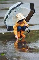 kind in geel waterbestendig mantel, laarzen en met paraplu spelen buitenshuis na de regen foto