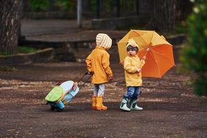 twee kinderen met paraplu, koffer en geel waterbestendig mantels en laarzen wandelen buitenshuis na de regen samen foto