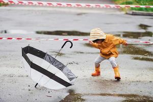 kind in geel waterbestendig mantel, laarzen en met paraplu spelen buitenshuis in de buurt beschermend plakband na de regen foto
