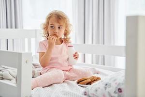 hebben een ontbijt door aan het eten koekjes. schattig weinig meisje in gewoontjes kleren is binnenshuis Bij huis Bij dag foto