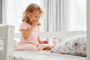 zittend Aan de bed, aan het eten koekjes en drinken melk. schattig weinig meisje in gewoontjes kleren is binnenshuis Bij huis Bij dag foto