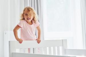 staand Aan de bed. schattig weinig meisje in gewoontjes kleren is binnenshuis Bij huis Bij dag foto