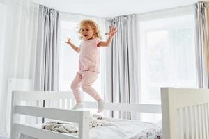 jumping Aan de bed. schattig weinig meisje in gewoontjes kleren is binnenshuis Bij huis Bij dag foto