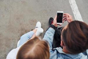 gebruik makend van telefoon. jong elegant Mens met vrouw in gewoontjes kleren zittend buitenshuis samen. opvatting van vriendschap of relaties foto