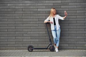 mooi blond in gewoontjes kleren staand met elektrisch schieter buitenshuis Bij zonnig dag met telefoon in handen foto