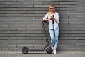 mooi blond in gewoontjes kleren staand met elektrisch schieter buitenshuis Bij zonnig dag met telefoon in handen foto