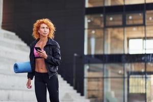 jong Europese roodharige vrouw in sportief kleren met telefoon en mat in handen buitenshuis foto