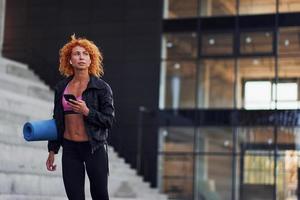 jong Europese roodharige vrouw in sportief kleren met telefoon en mat in handen buitenshuis foto