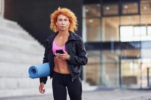 jong Europese roodharige vrouw in sportief kleren met telefoon en mat in handen buitenshuis foto