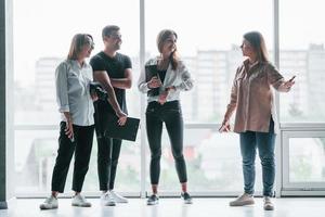 vrouw Holding haar professioneel camera. groep van bedrijf mensen in formeel kleren staand binnenshuis in de buurt groot venster foto