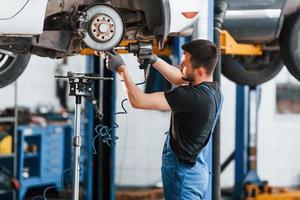 gebruik makend van speciaal hulpmiddel. Mens in werk uniform reparaties wit auto- binnenshuis. opvatting van auto onderhoud foto