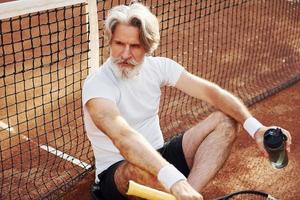 drinken water en zittend Aan de grond. senior modern elegant Mens met racket buitenshuis Aan tennis rechtbank Bij dag foto