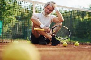 zit Aan de grond en duurt pauze. senior modern elegant Mens met racket buitenshuis Aan tennis rechtbank Bij dag foto