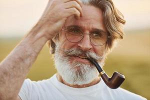 portret van senior elegant Mens met grijs haar- en baard dat staand buitenshuis Aan veld- Bij zonnig dag en roken foto