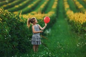 positief weinig meisje met rood ballon in handen hebben pret Aan de veld- Bij zomer dag tijd foto