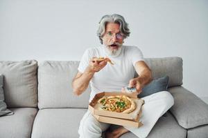 eet heerlijk pizza terwijl aan het kijken TV show. senior elegant modern Mens met grijs haar- en baard binnenshuis foto