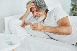 ziek senior elegant modern Mens met grijs haar- en baard binnenshuis aan het liegen Aan de bed en voelt slecht foto