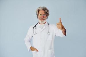 dokter in wit jas tegen muur. senior elegant modern Mens met grijs haar- en baard binnenshuis foto
