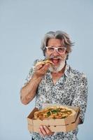 eet pizza wanneer staand tegen muur. senior elegant modern Mens met grijs haar- en baard binnenshuis foto