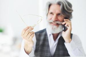geslaagd zakenman toepassingen telefoon. senior elegant modern Mens met grijs haar- en baard binnenshuis foto