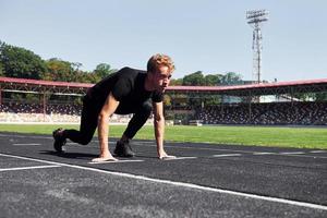 loper is Aan beginnend positie Aan spoor. sportief jong vent in zwart overhemd en broek buitenshuis Bij dag foto