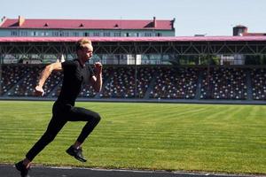 kant visie van loper Aan veld. sportief jong vent in zwart overhemd en broek buitenshuis Bij dag foto