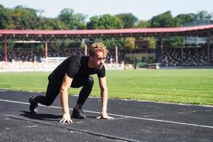 loper is Aan beginnend positie Aan spoor. sportief jong vent in zwart overhemd en broek buitenshuis Bij dag foto