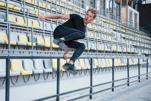 jumping en aan het doen parkour Aan de tribunes. sportief jong vent in zwart overhemd en broek buitenshuis Bij dag foto