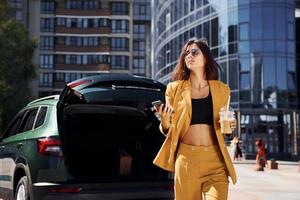 modern bedrijf gebouw Bij achtergrond. jong modieus vrouw in bordeaux gekleurde jas Bij dag met haar auto foto