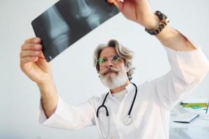 looks Bij röntgenfoto. senior mannetje dokter met grijs haar- en baard in wit jas is binnenshuis in kliniek foto