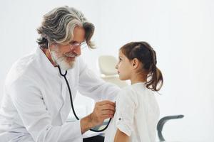 senior mannetje dokter met grijs haar- en baard in wit jas meten hart tarief van weinig meisje door stethoscoop binnenshuis in kliniek foto