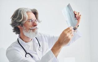 looks Bij röntgenfoto. senior mannetje dokter met grijs haar- en baard in wit jas is binnenshuis in kliniek foto