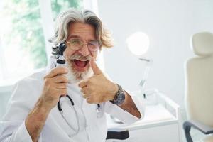 portret van senior mannetje dokter met grijs haar- en baard in wit jas is binnenshuis in otorinolaryngologie kliniek foto