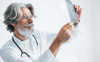 looks Bij röntgenfoto. senior mannetje dokter met grijs haar- en baard in wit jas is binnenshuis in kliniek foto